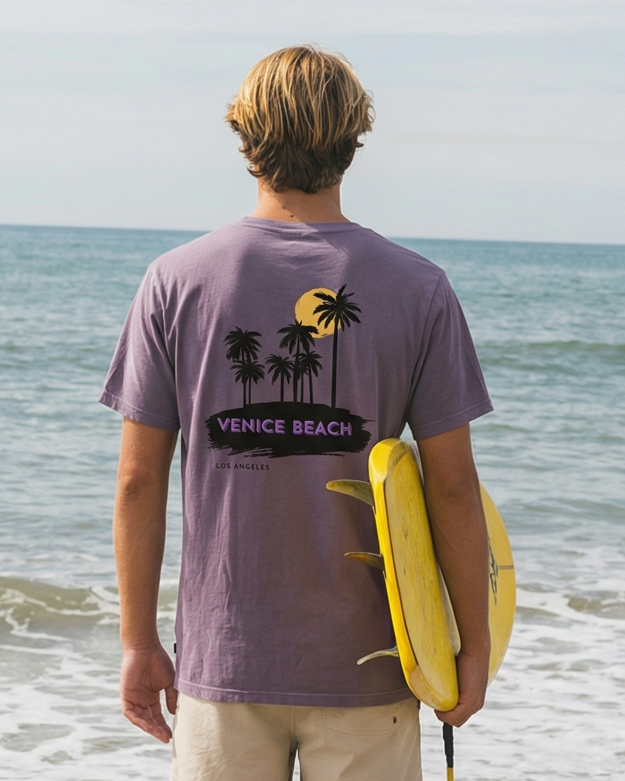Man standing on a beach holding a yellow surfboard, wearing a purple t-shirt with a palm tree design.
