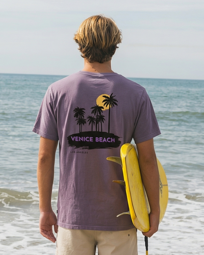 Man standing on a beach holding a yellow surfboard, wearing a purple t-shirt with a palm tree design.