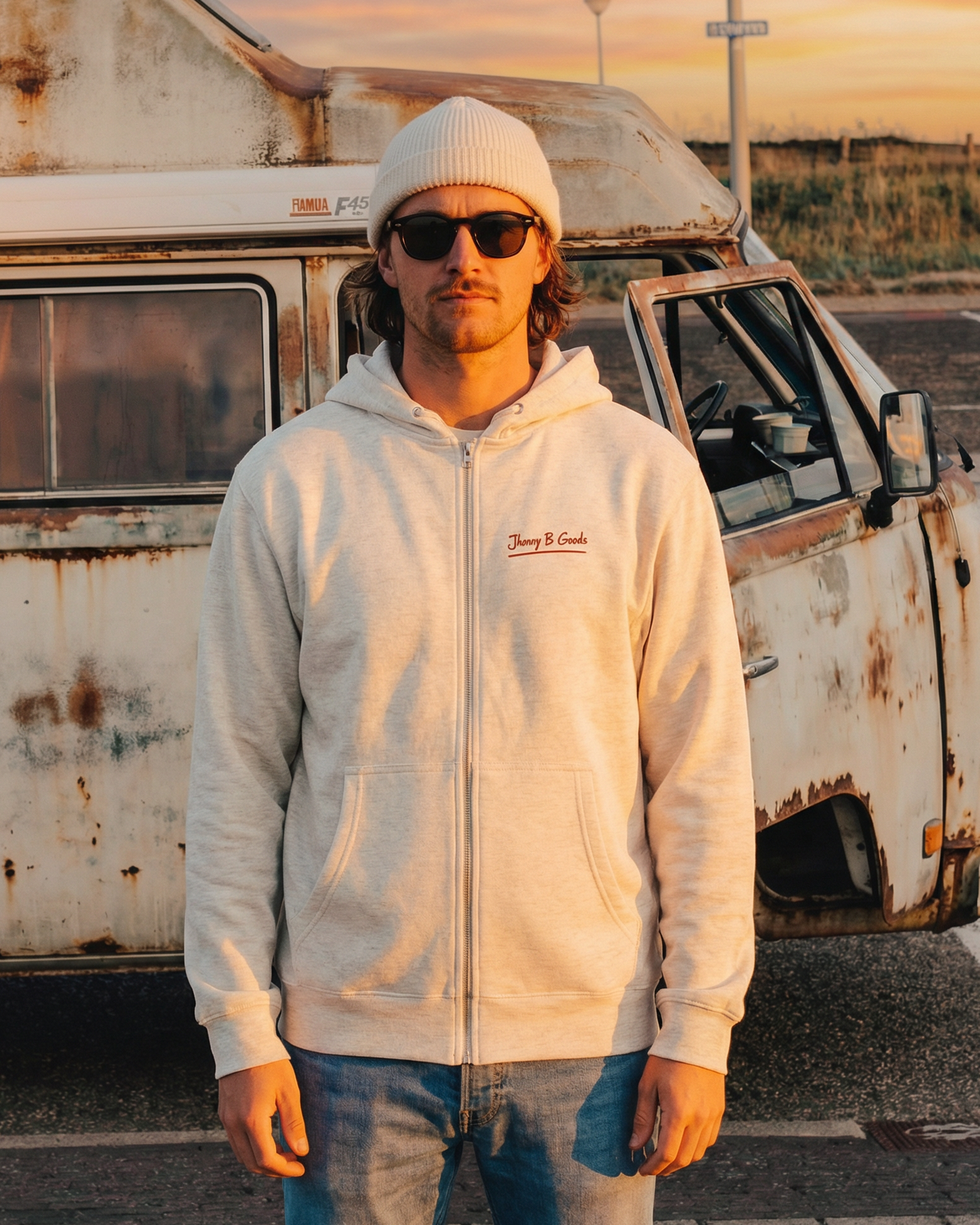Man wearing a white hoodie and sunglasses standing in front of a vintage van at sunset.