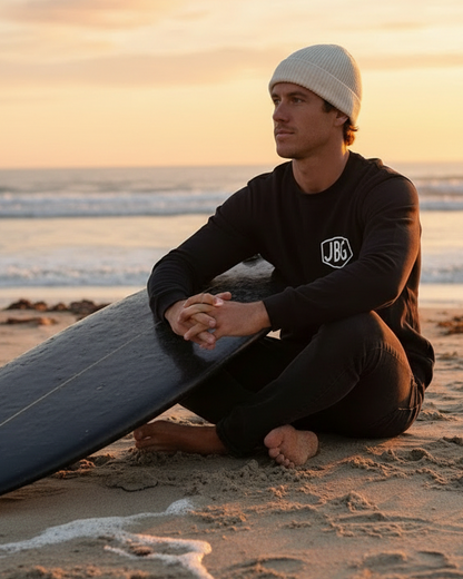 Person in a wetsuit sitting on the beach with a surfboard at sunset
