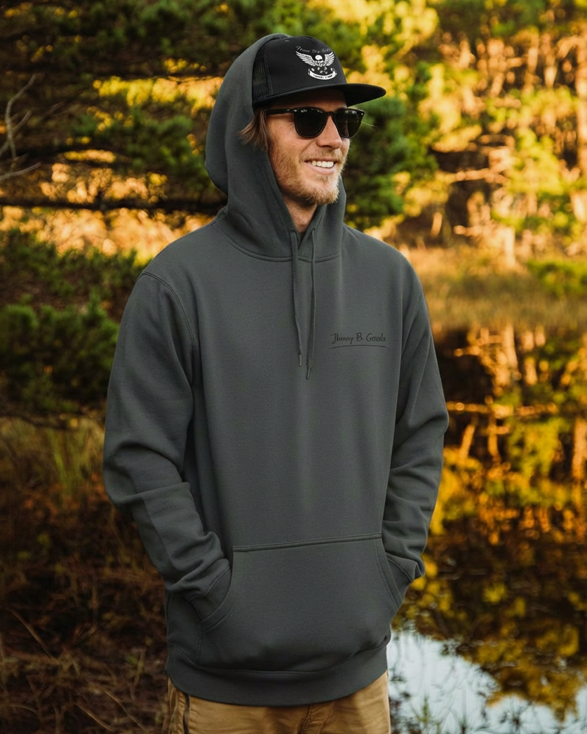 Person wearing a dark hoodie and cap standing by a body of water with trees in the background