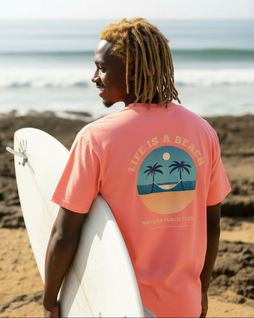 Person wearing a pink 'Life is a Beach' t-shirt holding a surfboard on a beach.