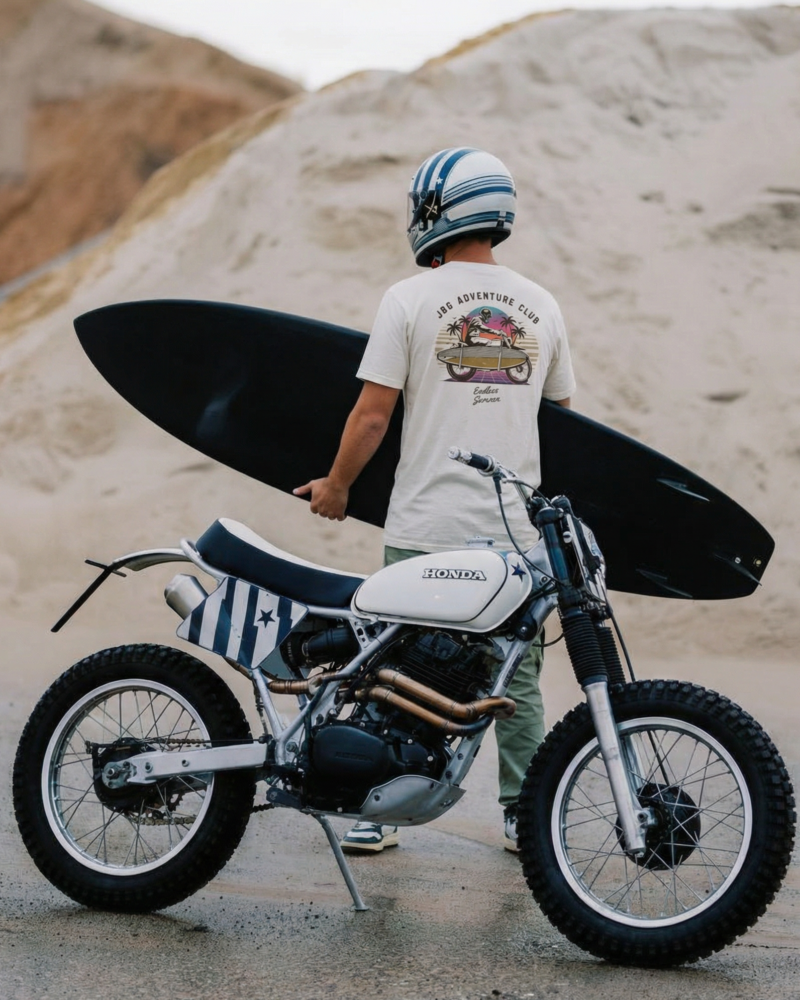 Person holding a surfboard standing next to a motorcycle on a sandy background