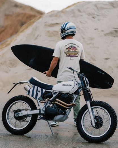 Person holding a surfboard standing next to a motorcycle on a sandy background