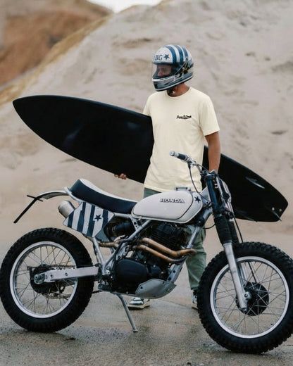 Person standing next to a motorcycle with a surfboard on a beach