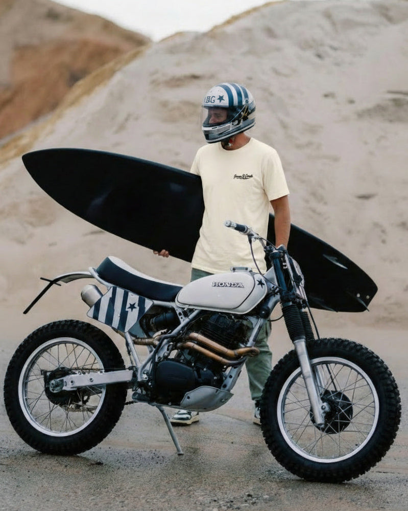 Person standing next to a motorcycle with a surfboard on a beach