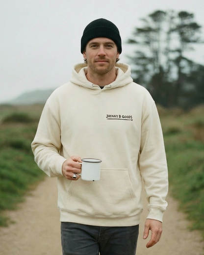 Man in a beige hoodie and black beanie holding a white cup on a path with grass and trees in the background