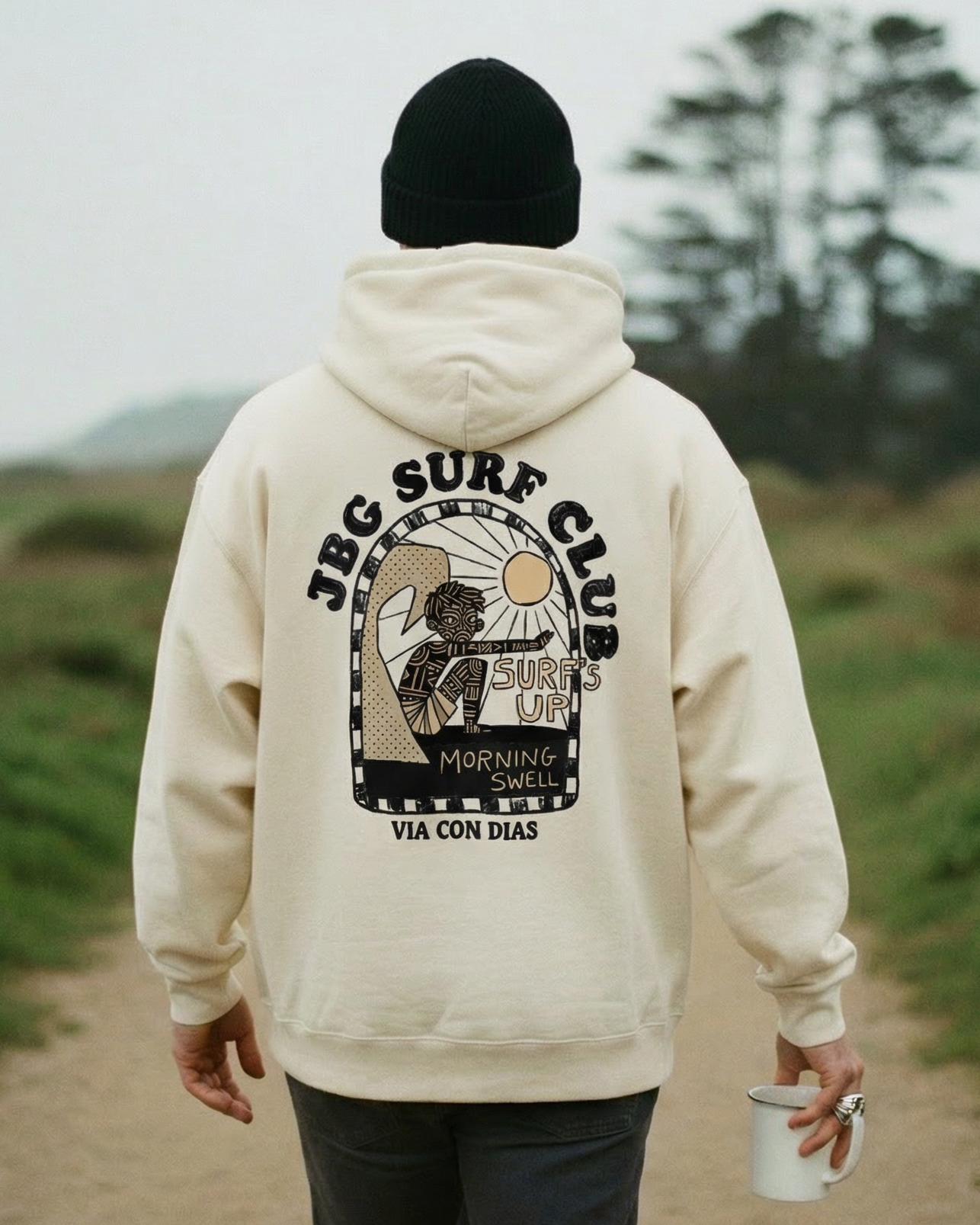 Person wearing a beige hoodie with a graphic design, walking on a path in a natural setting.