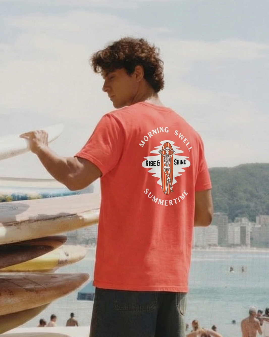Person in a red t-shirt with a graphic design, standing next to stacked surfboards on a beach.