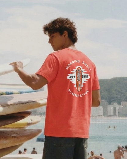 Person in a red t-shirt with a graphic design, standing next to stacked surfboards on a beach.