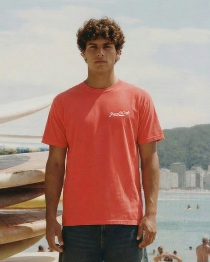 Man in a red t-shirt and denim shorts standing on a beach with surfboards stacked behind him.