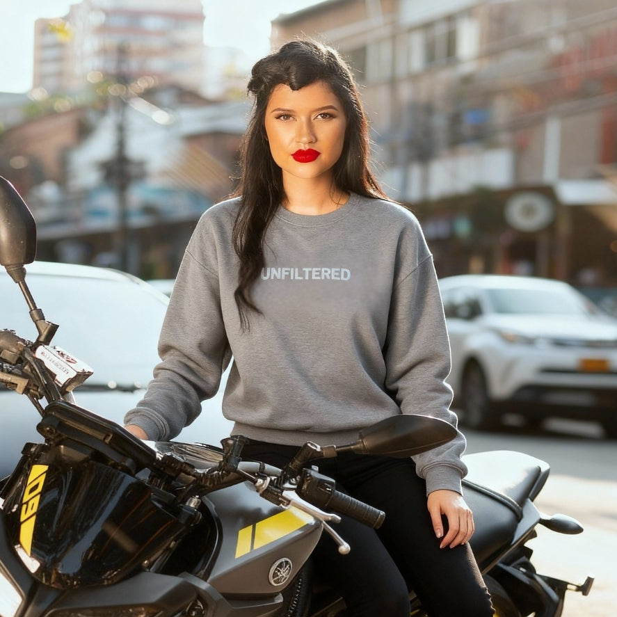Woman wearing a gray sweatshirt with 'UNFILTERED' text, sitting on a motorcycle in an urban setting.