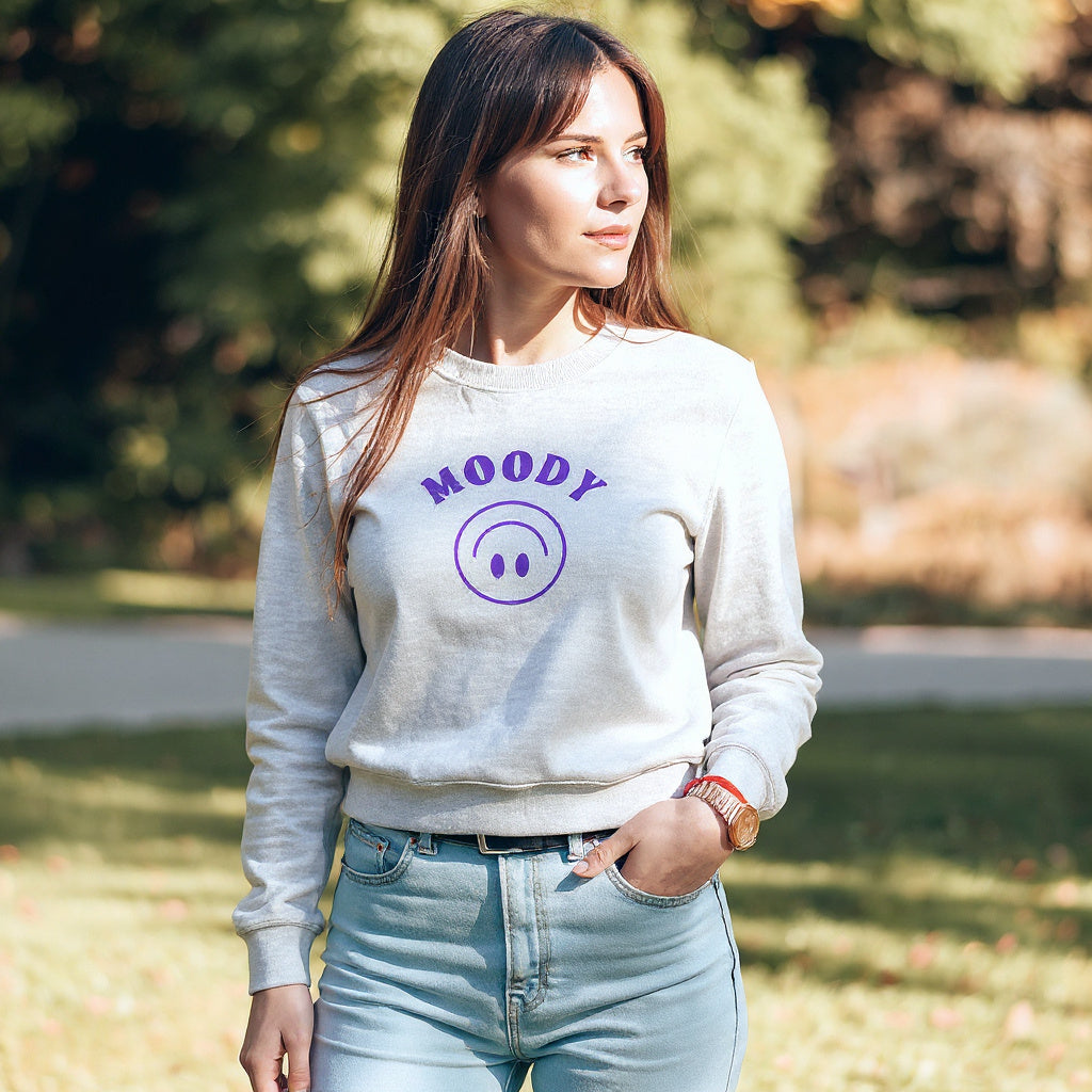 Woman wearing a light gray JBG sweatshirt with Embroidered 'MOODY' and a purple circle design, standing outdoors.d 