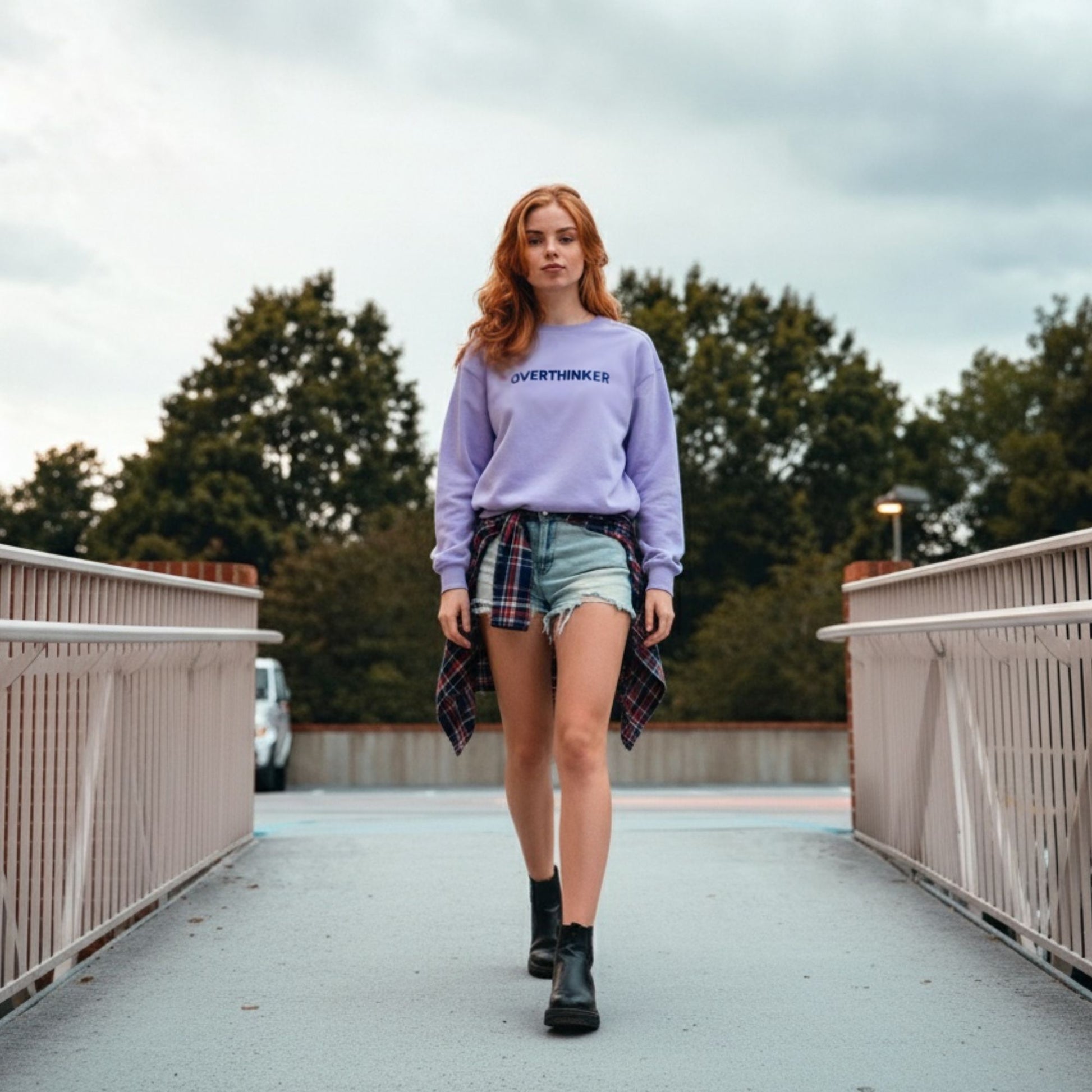 Woman wearing a lavender sweatshirt with text, plaid shirt, and denim shorts on a bridge.