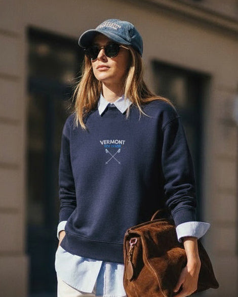 Woman wearing a blue sweatshirt with 'Vermont' text, holding a brown bag on a city street.