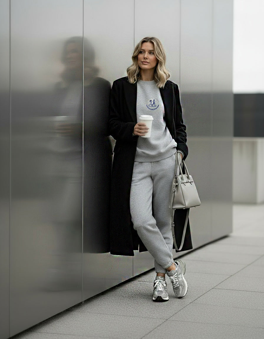 Woman in a gray tracksuit with a black coat leaning against a metallic wall.