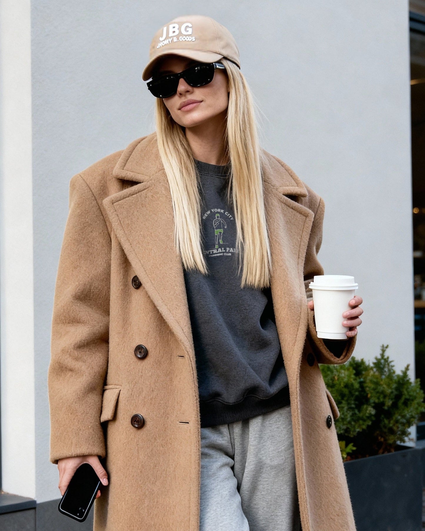 Person wearing a beige coat and cap, holding a coffee cup, standing outside a building.