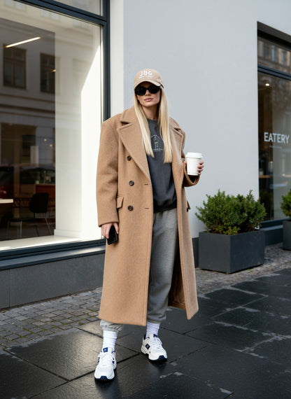 Person wearing a long beige coat and sunglasses, holding a coffee cup on a city street.