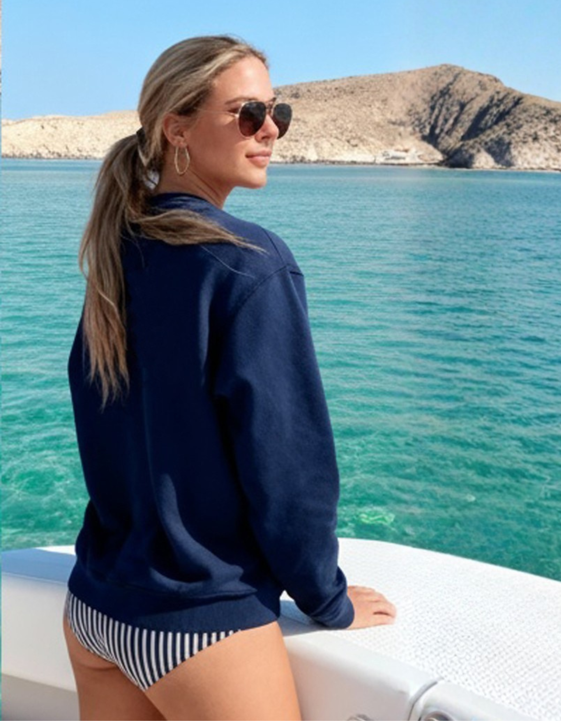 Woman in a navy jacket and striped swimsuit on a boat with a scenic background