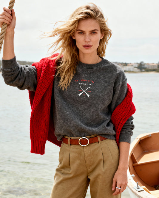Woman standing by a boat on a beach wearing a gray sweatshirt with a red cardigan and beige pants.