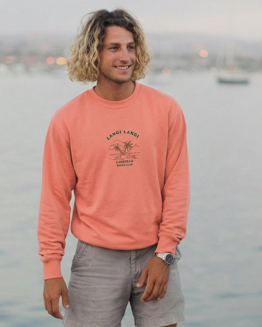 Man wearing a pink long-sleeve shirt and gray shorts standing on a dock with water and boats in the background.