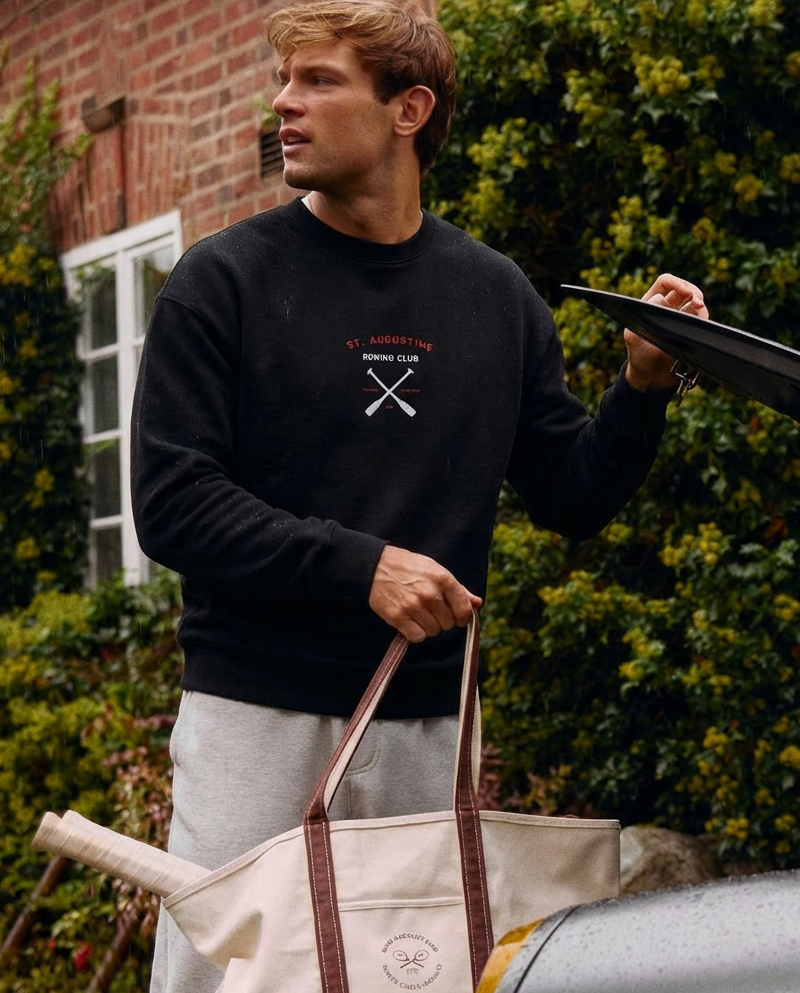Man holding a black umbrella and a beige tote bag with a car and greenery in the background