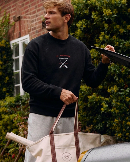 Man holding a black umbrella and a beige tote bag with a car and greenery in the background