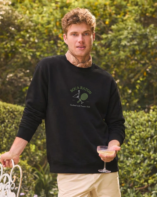 Man wearing a black sweatshirt with a logo, holding a drink outdoors.
