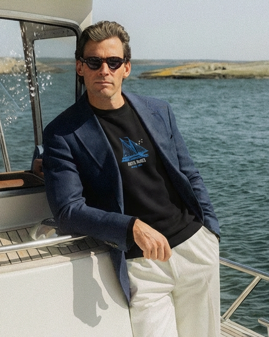 Man on a boat wearing sunglasses and a jacket with a logo, with water and land in the background.