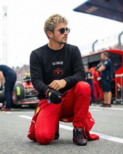 Person in red racing suit and black shirt sitting on a race track with pit crew in the background