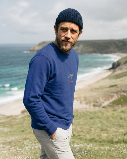 Man wearing a blue sweater and knit cap standing on a grassy area with ocean and cliffs in the background