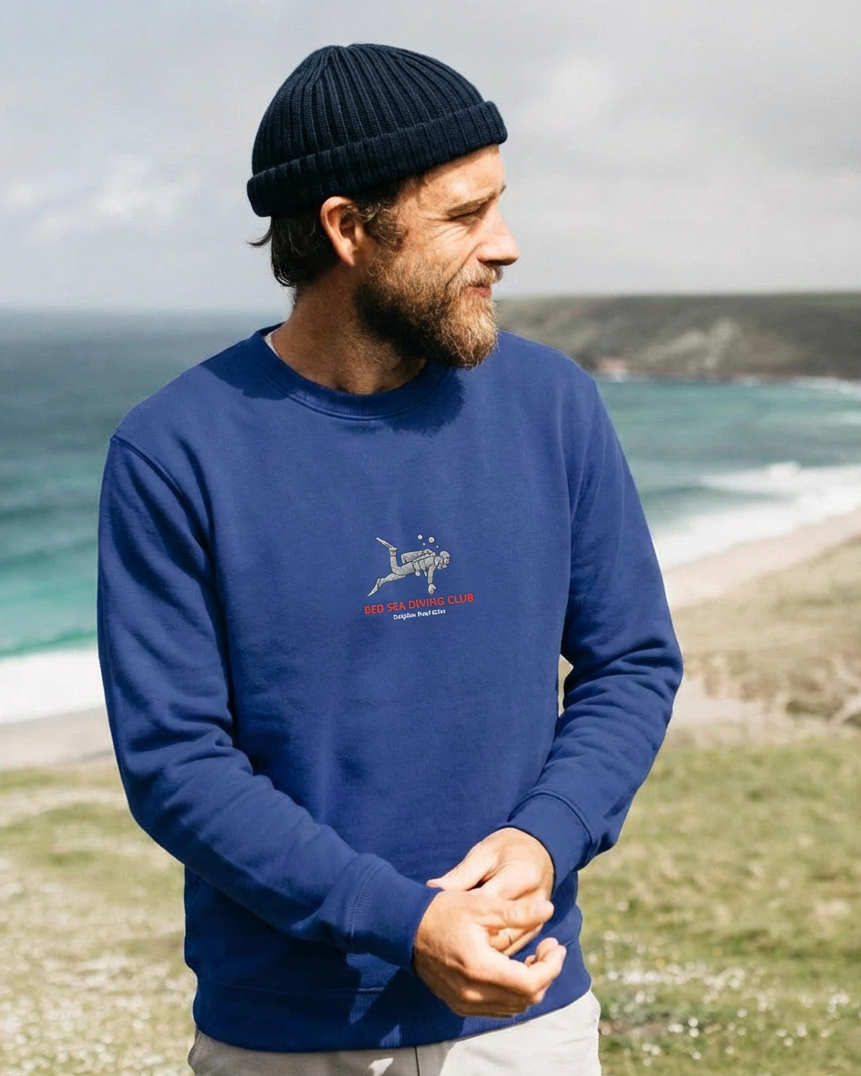 Man wearing a blue sweatshirt with a logo standing by the ocean.