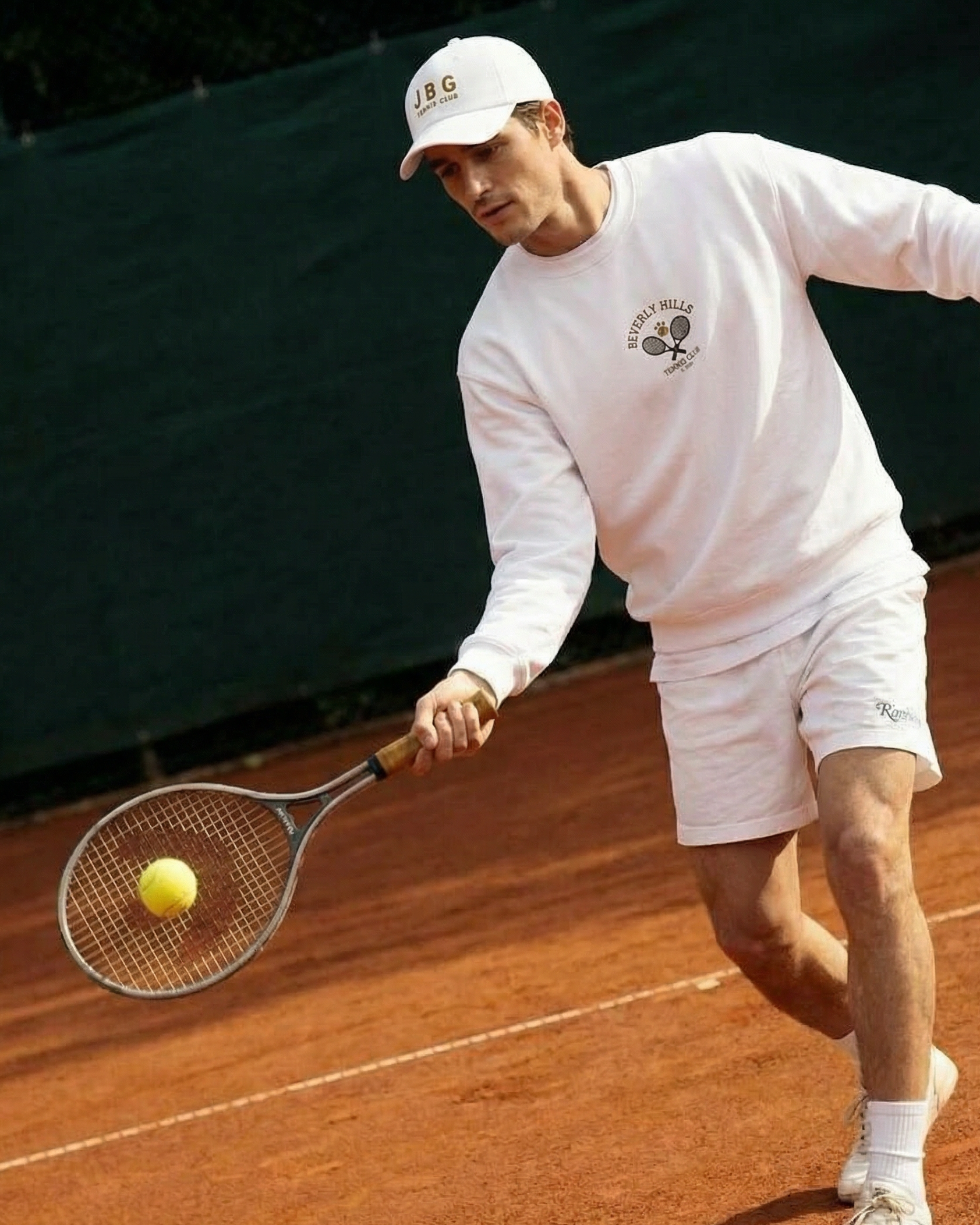 Man playing tennis on a clay court, wearing a white outfit and cap.
