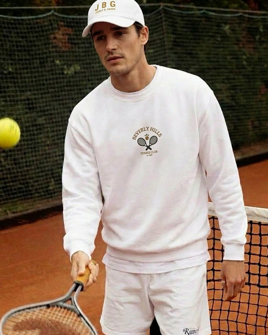 Man on a tennis court holding a racket and a ball, wearing a white sweatshirt and shorts.