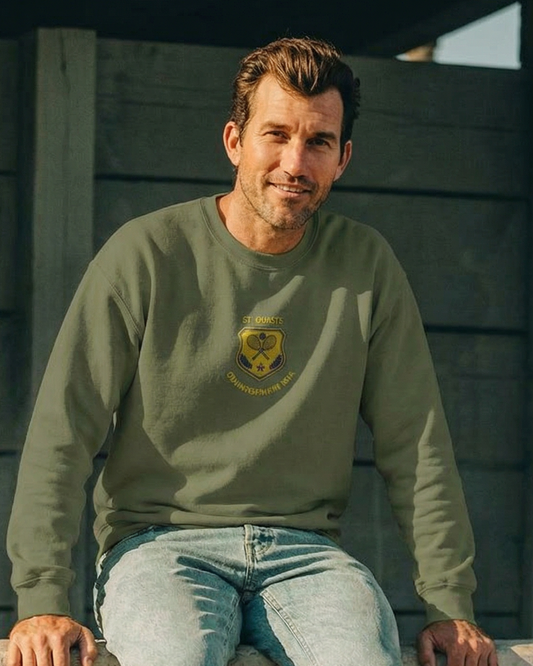 Man wearing a green sweatshirt with a logo and light blue jeans sitting on a concrete ledge.