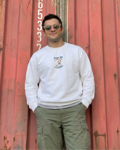 Man wearing a white sweatshirt with a logo and sunglasses, standing in front of a red shipping container.
