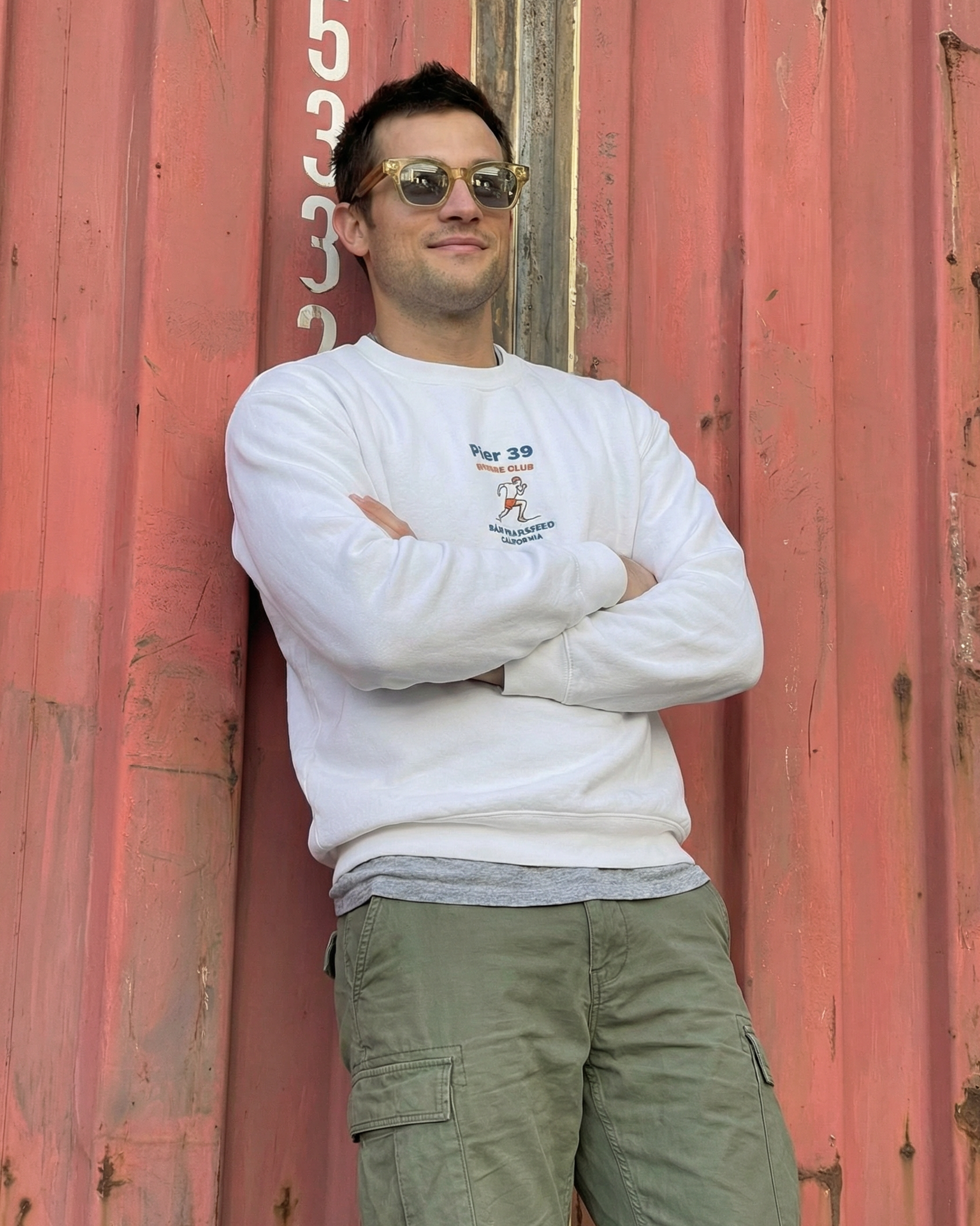 Man wearing a white sweatshirt and green pants standing against a red metal wall.