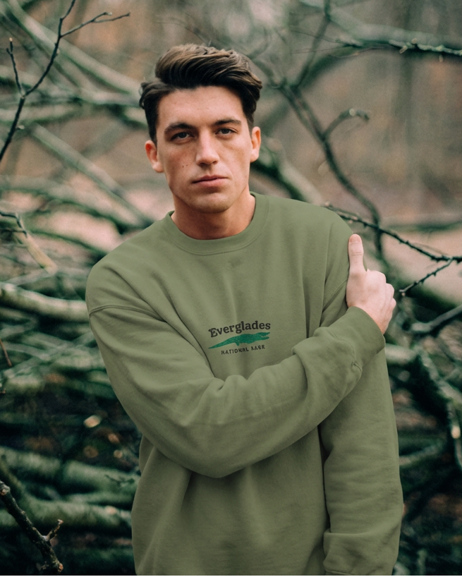 Man wearing a green sweatshirt with a logo, standing among bare tree branches.