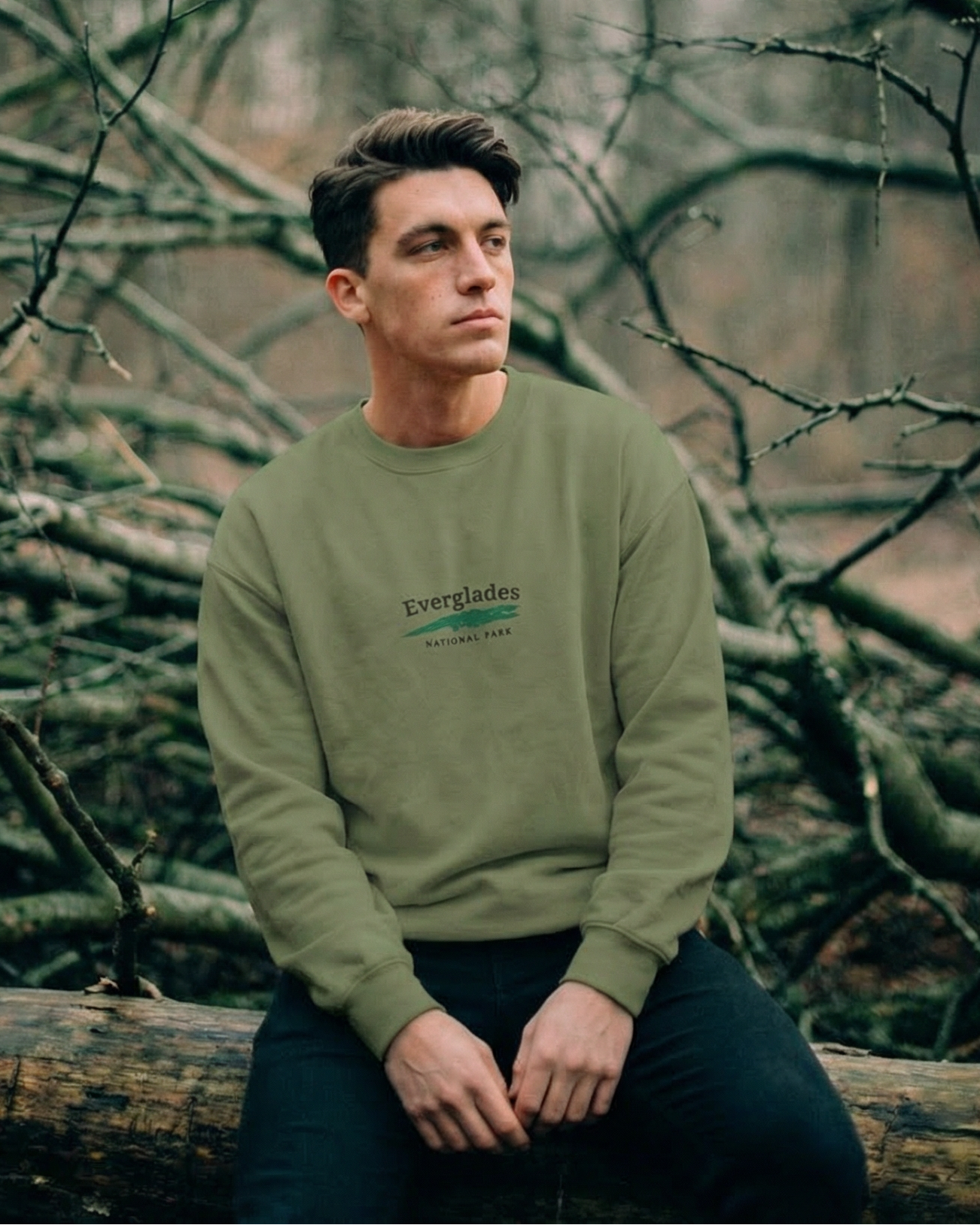 Man sitting on a log in a forest wearing a green sweatshirt with text.