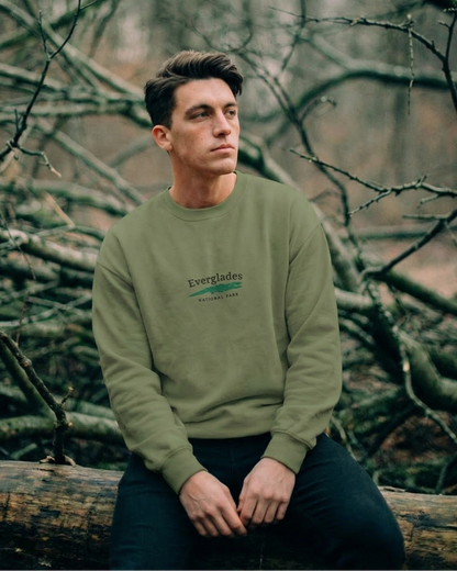 Man sitting on a log in a forest wearing a green sweatshirt with text.