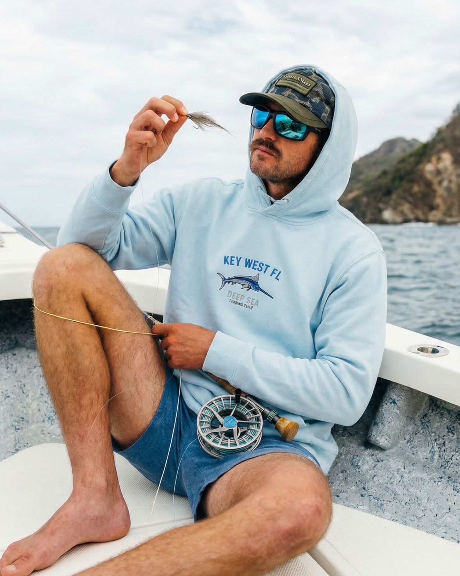Man on a boat holding a fish, wearing a 'Key West FL' hoodie and sunglasses.