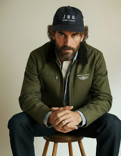 Man wearing a green jacket and black cap with 'JBG' logo, sitting on a stool against a plain background.