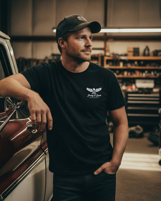 Man leaning against a classic car in a garage wearing a black t-shirt with a logo.