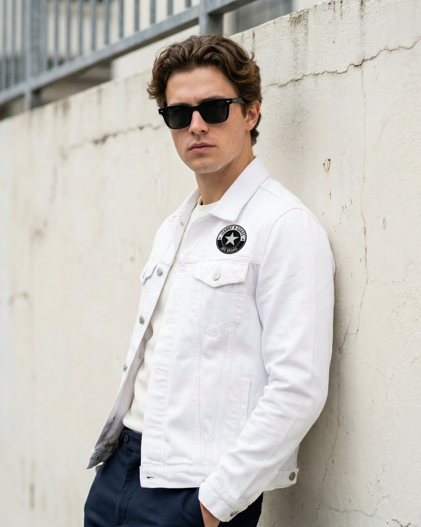 Man wearing a white jacket with a logo, sunglasses, and dark pants against a concrete wall.