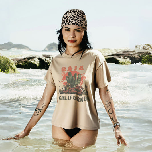 A woman standing in the ocean wearing a beige oversized t-shirt with 'Baja California' text and a graphic of a cactus, snake, and leopard, paired with black cutoff shorts.