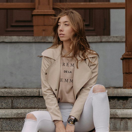 A woman sitting on steps wearing a beige jacket and a beige t-shirt with text on it, paired with white pants.