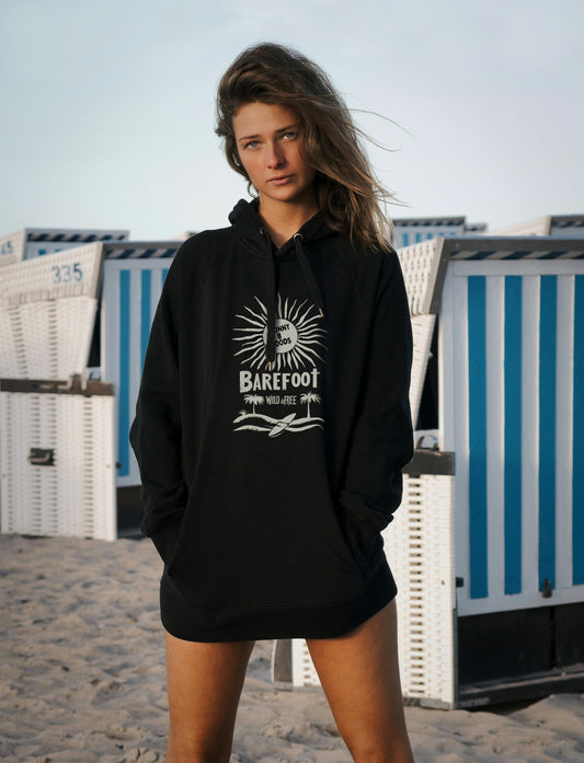 A woman standing on a beach wearing a black hoodie with a graphic print and the text 'Barefoot Wild & Free' on the center chest.