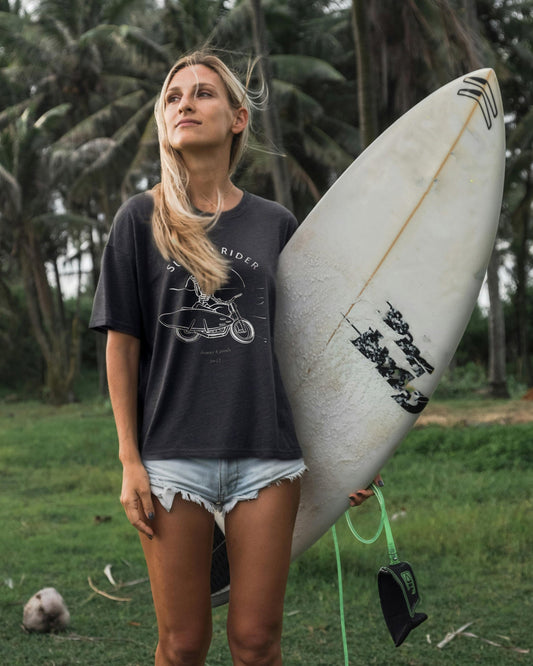A woman standing with a surfboard wearing a black t-shirt with a graphic of a skeleton riding a motorcycle, paired with denim shorts.