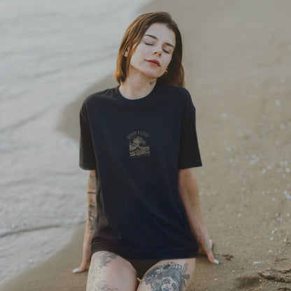 A woman sitting on the beach wearing a black crew neck t-shirt with a retro wave design and the text 'Pacific Wave' and 'Santa Cruz, California' printed on it.