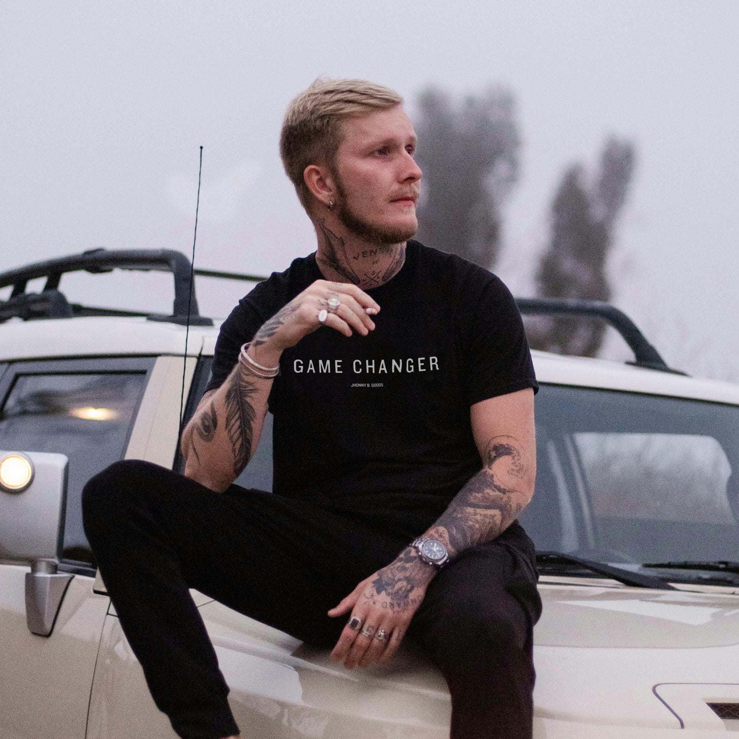 A man sitting on a car bonnet wearing a black t-shirt with 'GAME CHANGER' text in white.